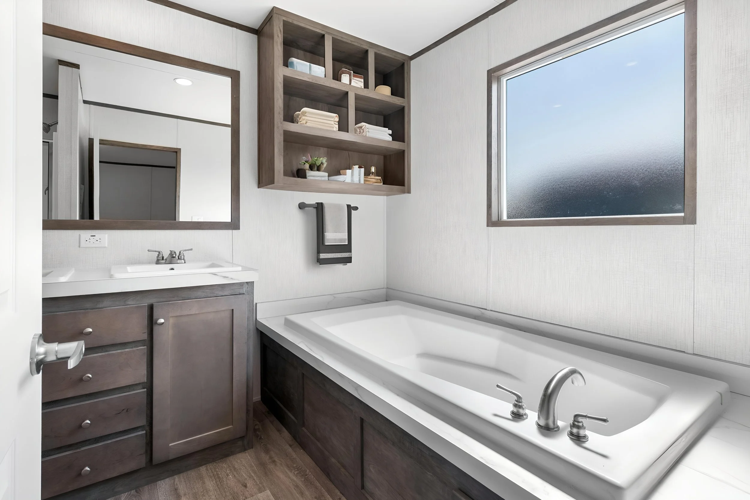 A bathroom with a bathtub, a sink with a mirror, a wall-mounted shelf with towels and decor, and a frosted window.