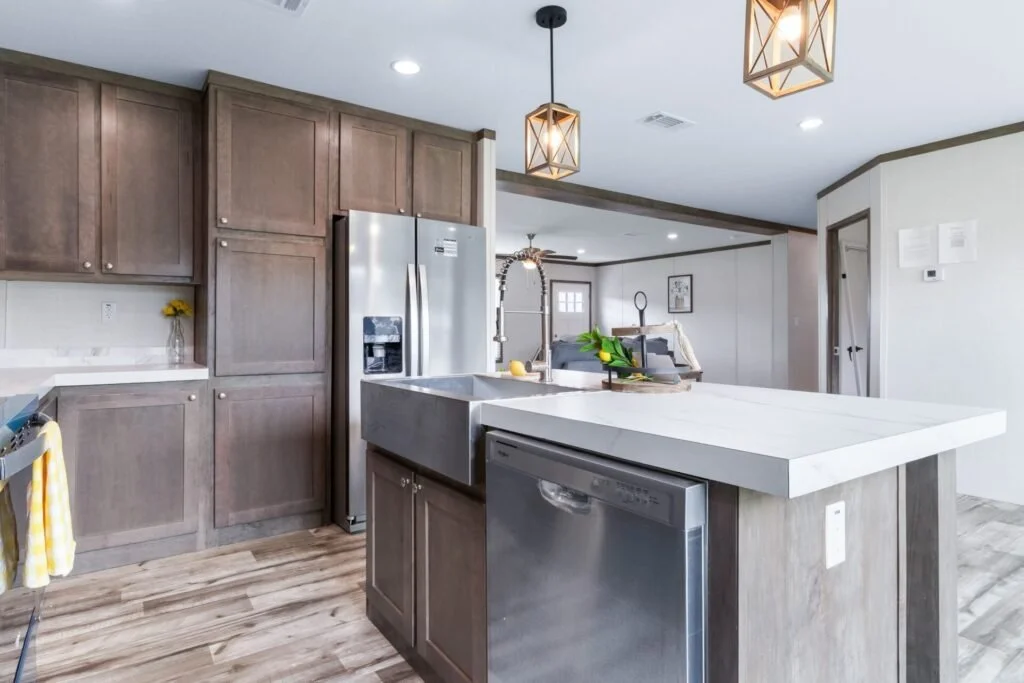 Modern kitchen with gray cabinets, stainless steel refrigerator, and white countertops. Pendant lights hang above a kitchen island with a farmhouse sink. The open layout connects to a living area with a sofa and artwork.