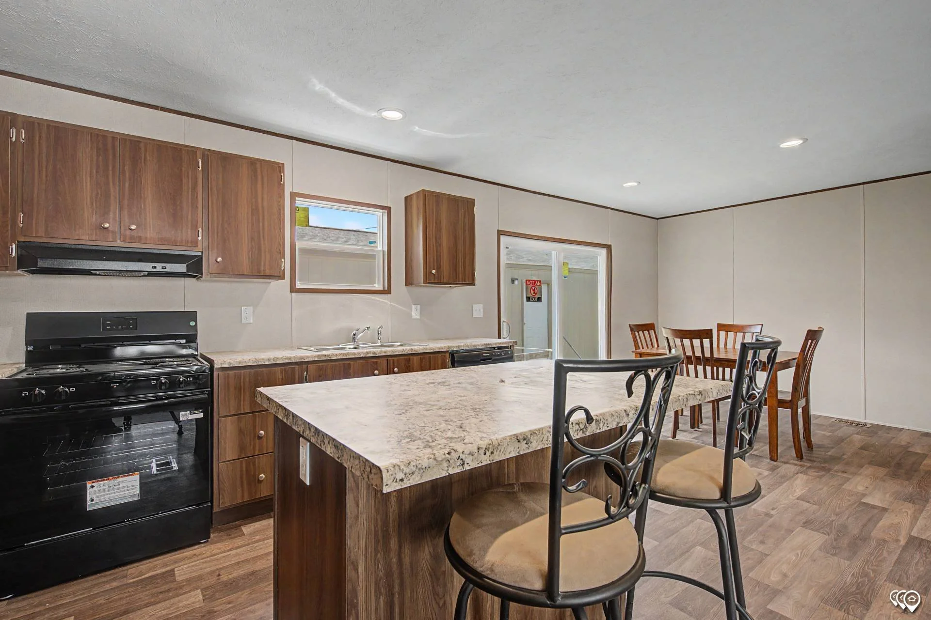 A kitchen with dark wood cabinets, a black oven, a beige speckled countertop island with black metal chairs, and a dining area with a wooden table and chairs. There are two small windows and a door in the kitchen area.