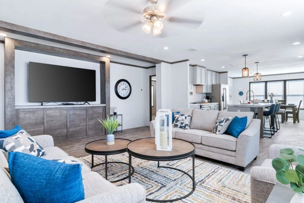 Open-concept living room and kitchen with modern furniture, a large flat-screen TV, a clock on the wall, and a ceiling fan.