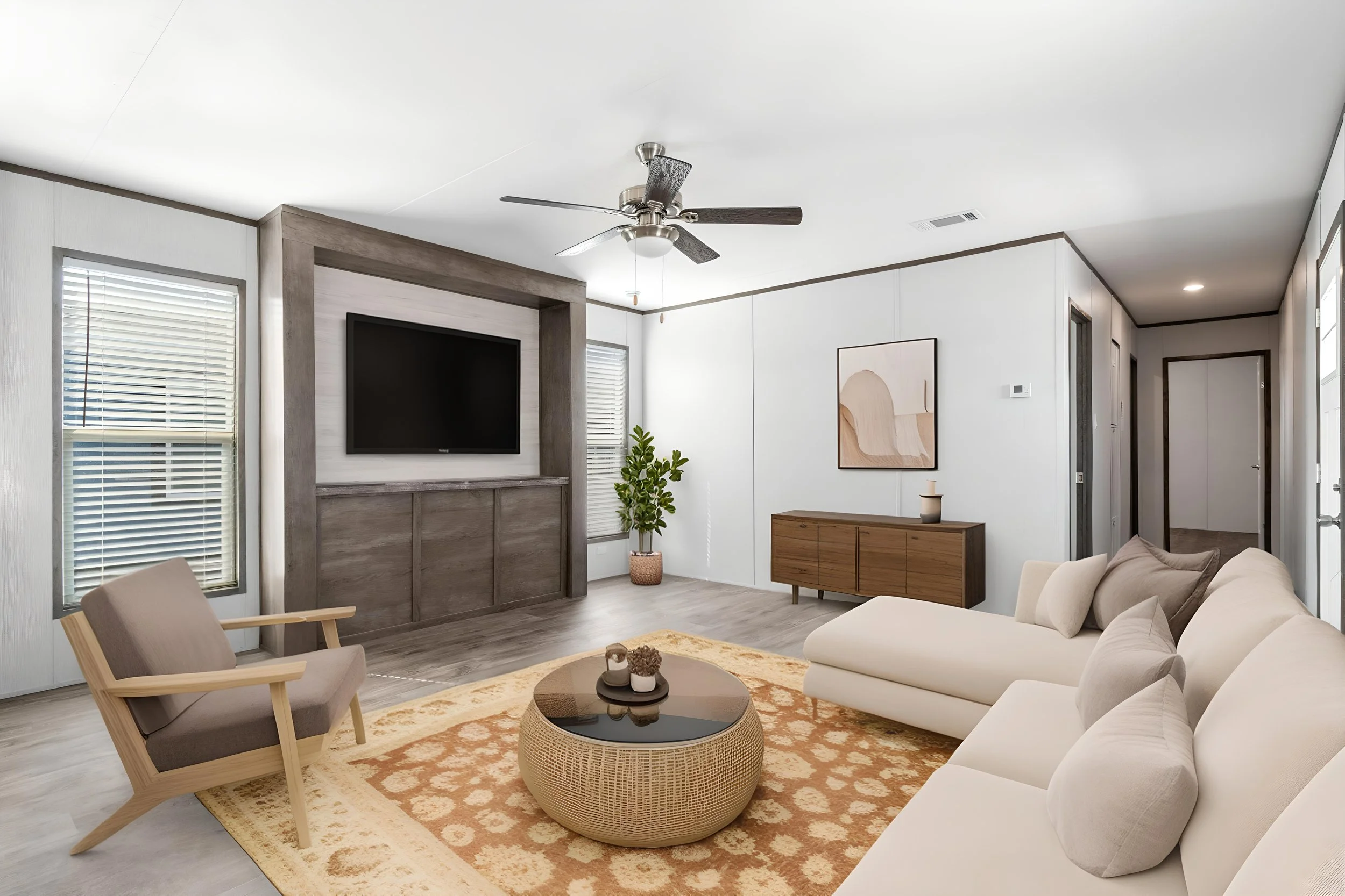 Living room with white walls, beige sofa, beige armchair, wooden coffee table, patterned rug, mounted flat-screen TV, window with blinds, potted plant, centered ceiling fan, abstract artwork, and additional furniture along the wall.