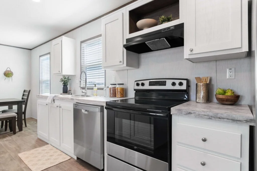 Modern kitchen with white cabinets, a black oven, a stainless steel dishwasher, and a marble countertop. There are two windows with blinds, a sink under one window, decorative items on the counter, and a small dining table with chairs in the backgrou