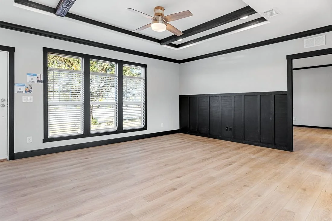 Empty living room with large windows, light wood flooring, white walls with black trim, and a ceiling fan with wooden blades.