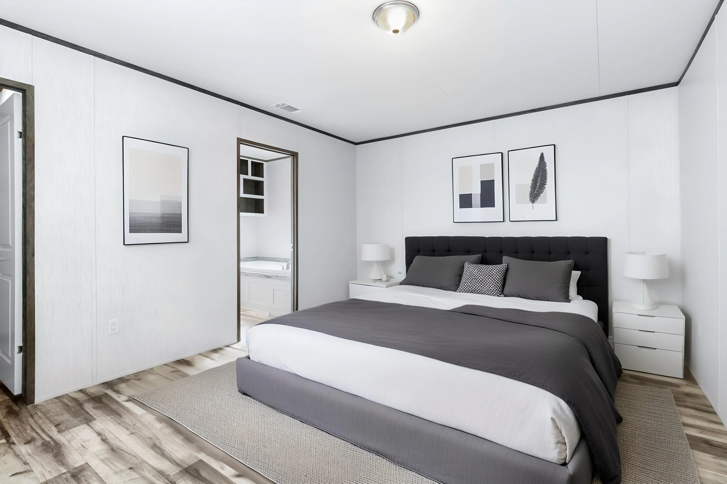 A bedroom with a neatly made bed featuring dark gray bedding, white pillows, and black-and-white accent pillows. The room has white walls with black trim, modern artwork above the bed, and two white nightstands with lamps. There's a small window open