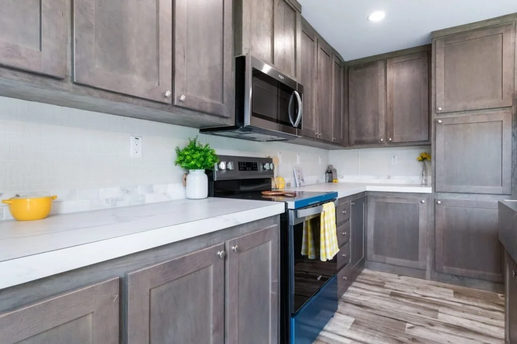 Modern kitchen with gray cabinets, white countertops, stainless steel microwave and oven, and wooden flooring. Decor includes green plant, yellow bowl, and small vase with yellow flowers.