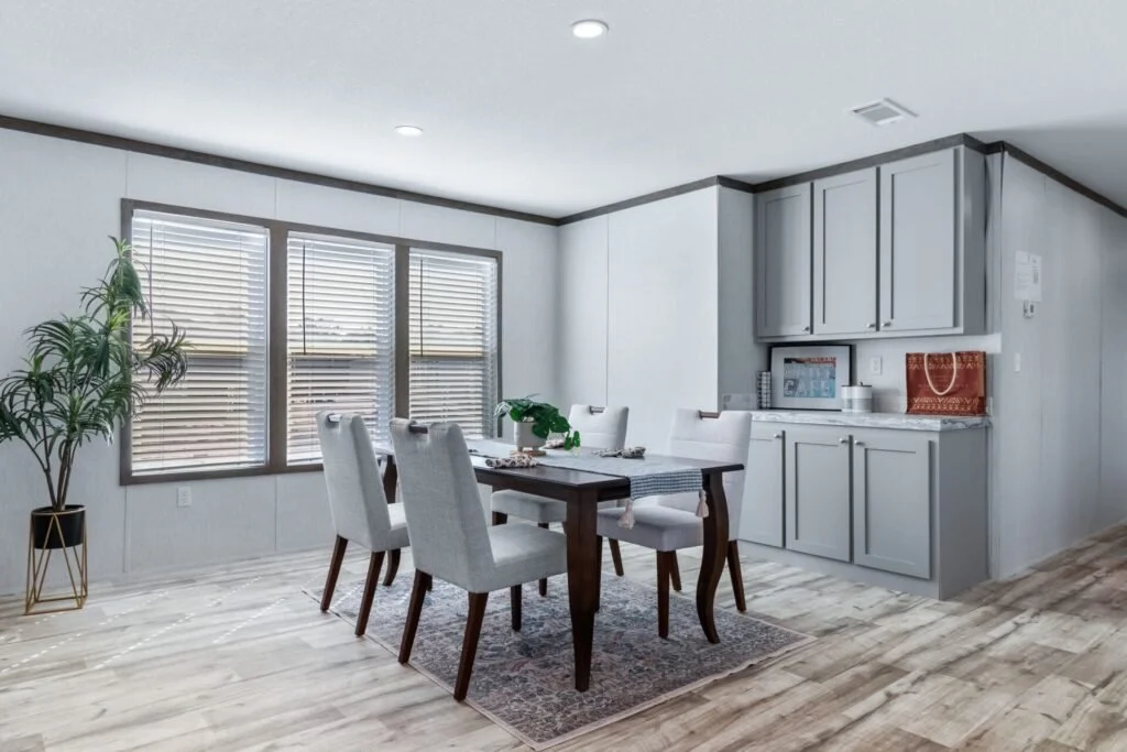A dining room with a wooden table, six white upholstered chairs, and a sideboard with gray cabinets. There is a potted plant in a black and gold stand near the window with blinds. The floor is light-colored wood, and the walls are painted light gray.