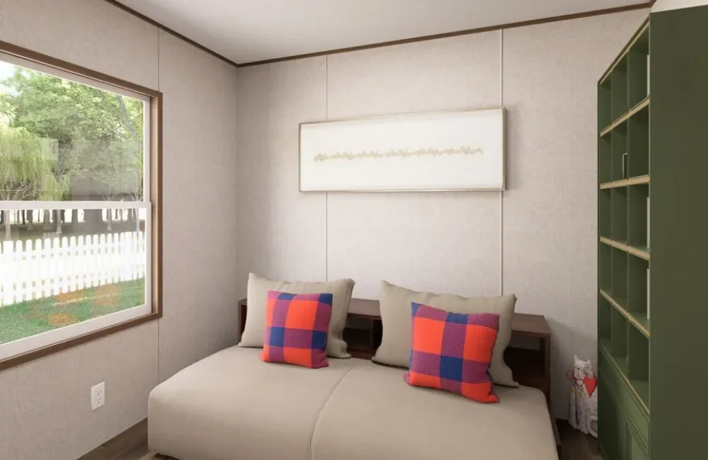 A small bedroom with a window showing a white picket fence and greenery outside. The room has light-colored walls, a minimalist art piece, and a green bookshelf on the right. There is a bed with two beige pillows and two colorful plaid pillows.