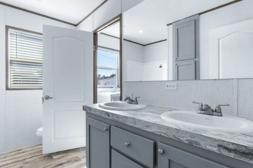 Bathroom with double sink vanity, marble countertop, blue cabinets, mirror, window with blinds, open door, and glimpse of toilet and outdoor view.