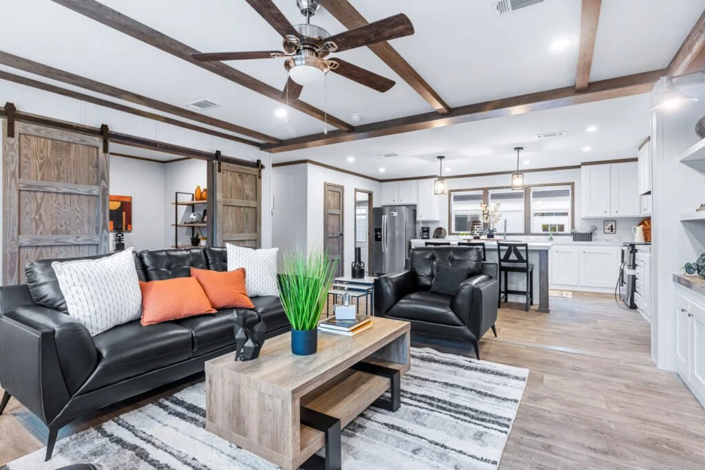 Living room with black leather sofas, wooden coffee table with a green plant, striped rug, open kitchen with white cabinets, stainless steel refrigerator, and hanging pendant lights.