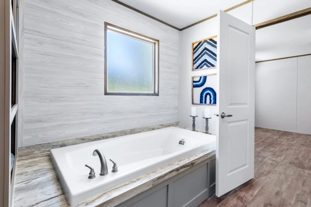 Bathroom with a white bathtub, a window above it, and two candles on candelabras beside the open door. Framed artwork on the wall, and wood floors.