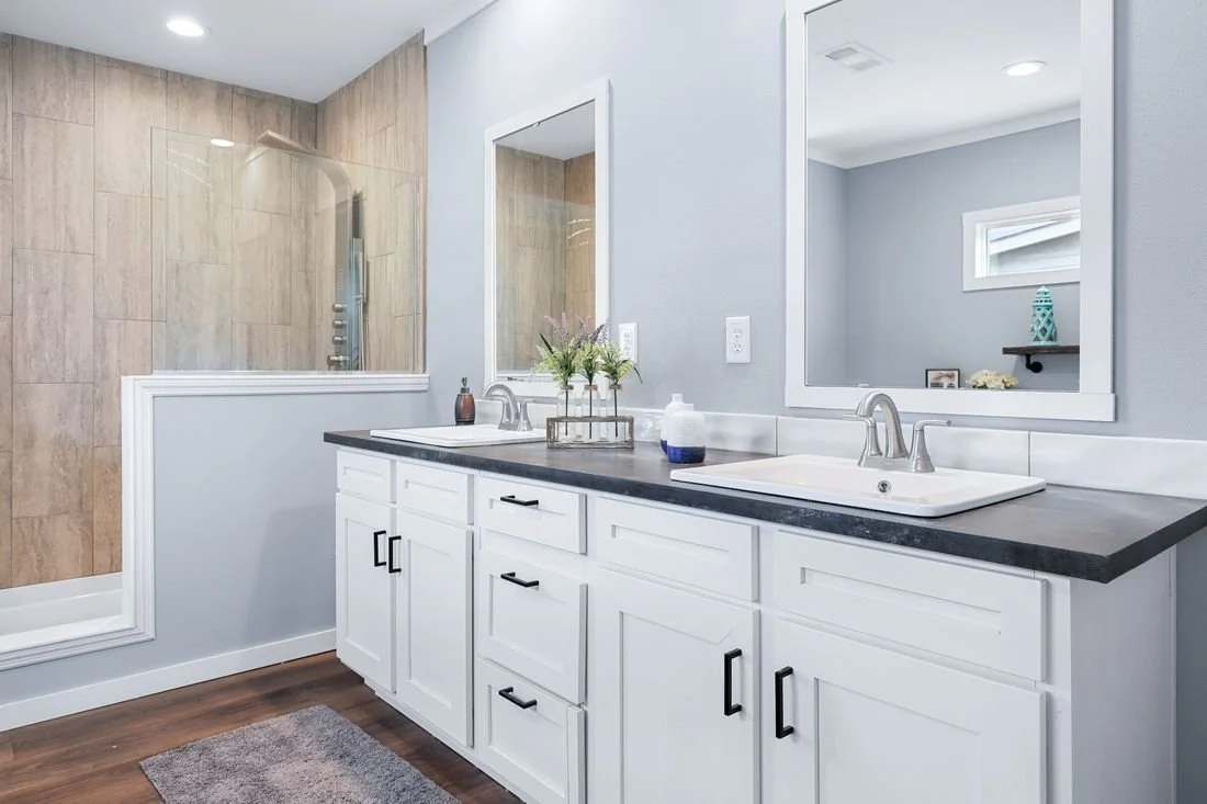 Bathroom with double vanity, black countertop, two sinks, large mirror, light gray walls, shower with glass door and tan tiles, small window, decorative items, wooden floor, gray bath mat.