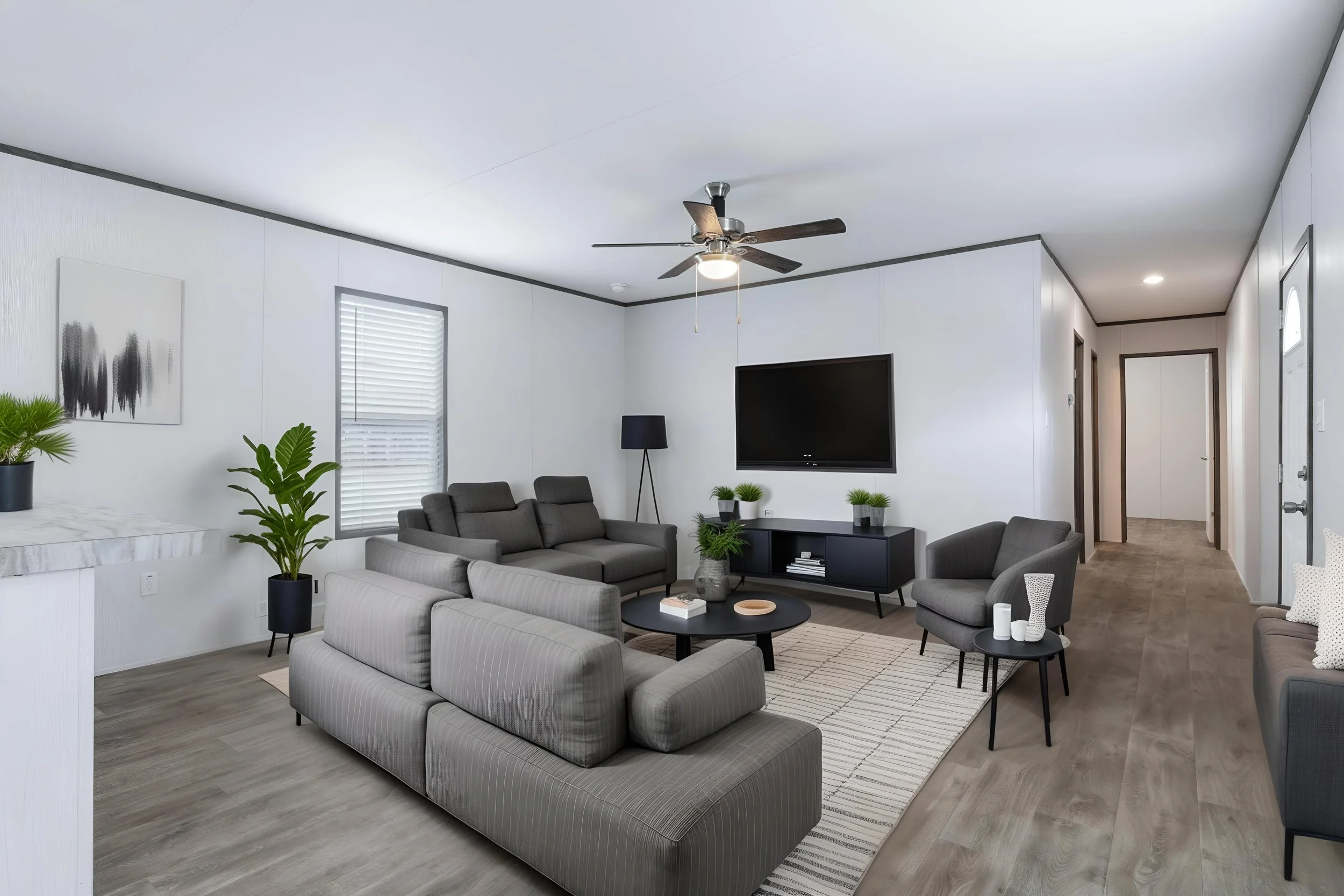 Modern living room with gray sofas, black coffee table, plants, wall art, flat-screen TV, floor lamp, ceiling fan, and hallway with doors.