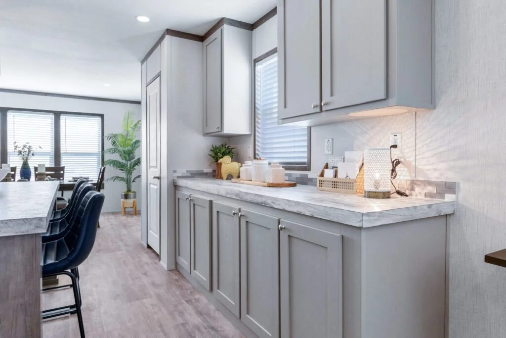 Kitchen with light gray cabinets, marble countertop, window with blinds, and decorative items including a potted plant and lamps, with a view of a dining area and large windows in the background.