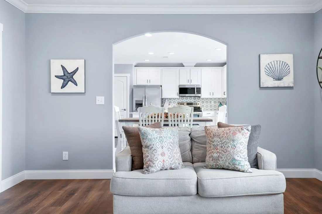 Living room with a beige sofa and decorative pillows, wall art with a starfish and a shell, and an open view to a kitchen with white cabinets and stainless steel appliances.