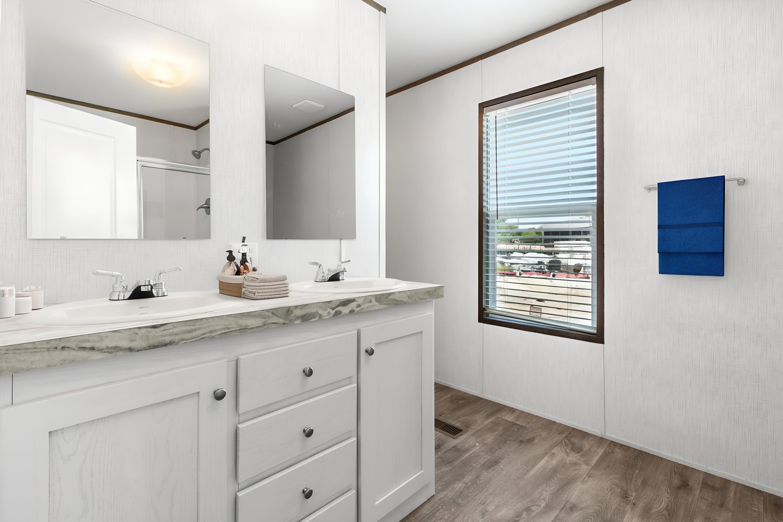 Clean bathroom with white vanity, marble countertop, two sinks, large mirror, window with blinds, and blue towel hanging on wall.