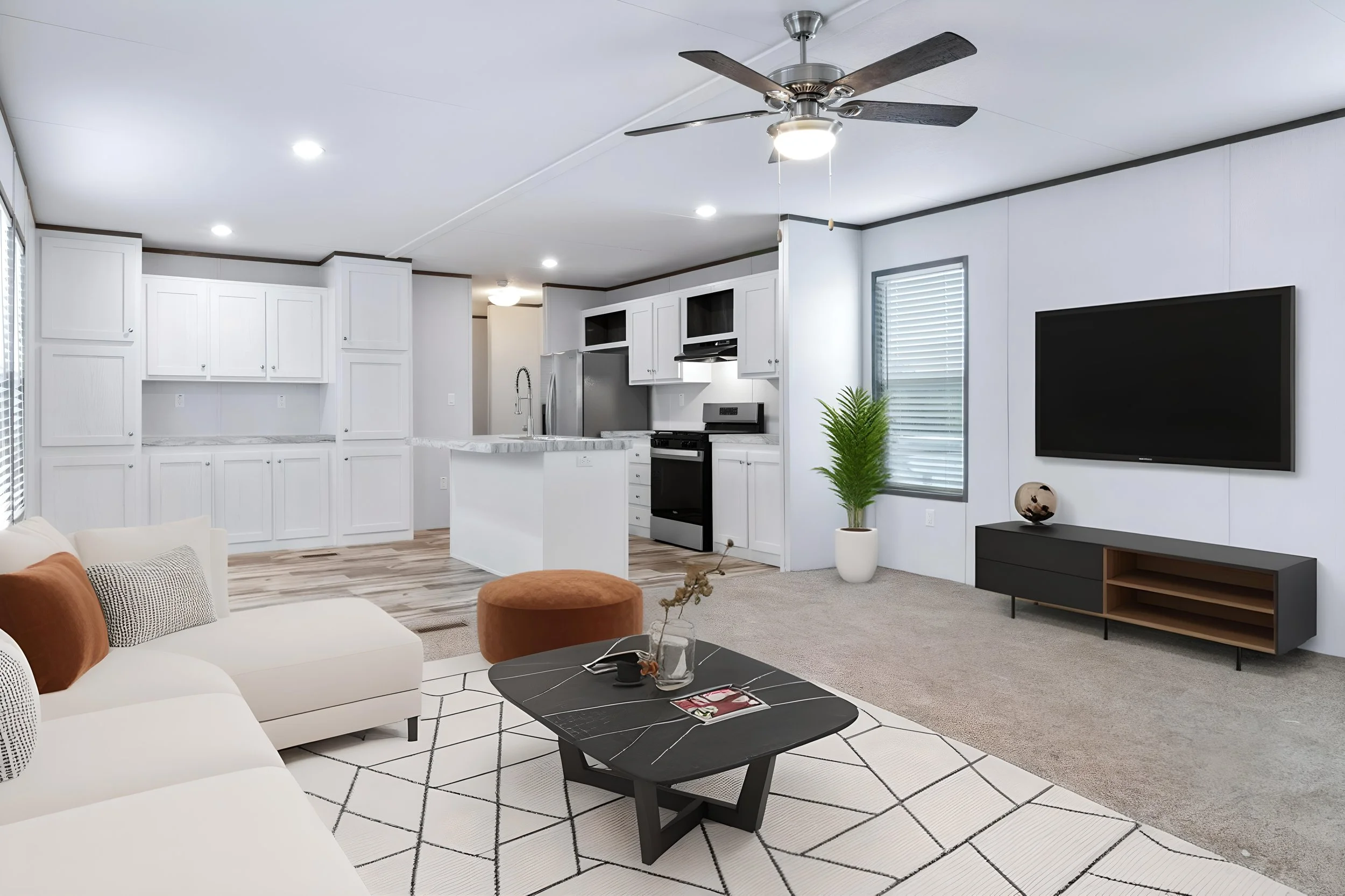Living room with white sofa, black coffee table, orange ottoman, and geometric area rug. Kitchen in the background with white cabinetry, marble countertops, and stainless steel appliances. Large flat-screen TV on the wall, with a potted plant and decor on a black media console. Ceiling fan and windows with blinds provide natural light.