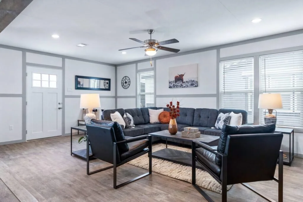Living room with a large gray sectional sofa, two black leather armchairs, and a coffee table with a vase of red flowers, beige rug, and lamps on side tables. Bright with windows and a ceiling fan.