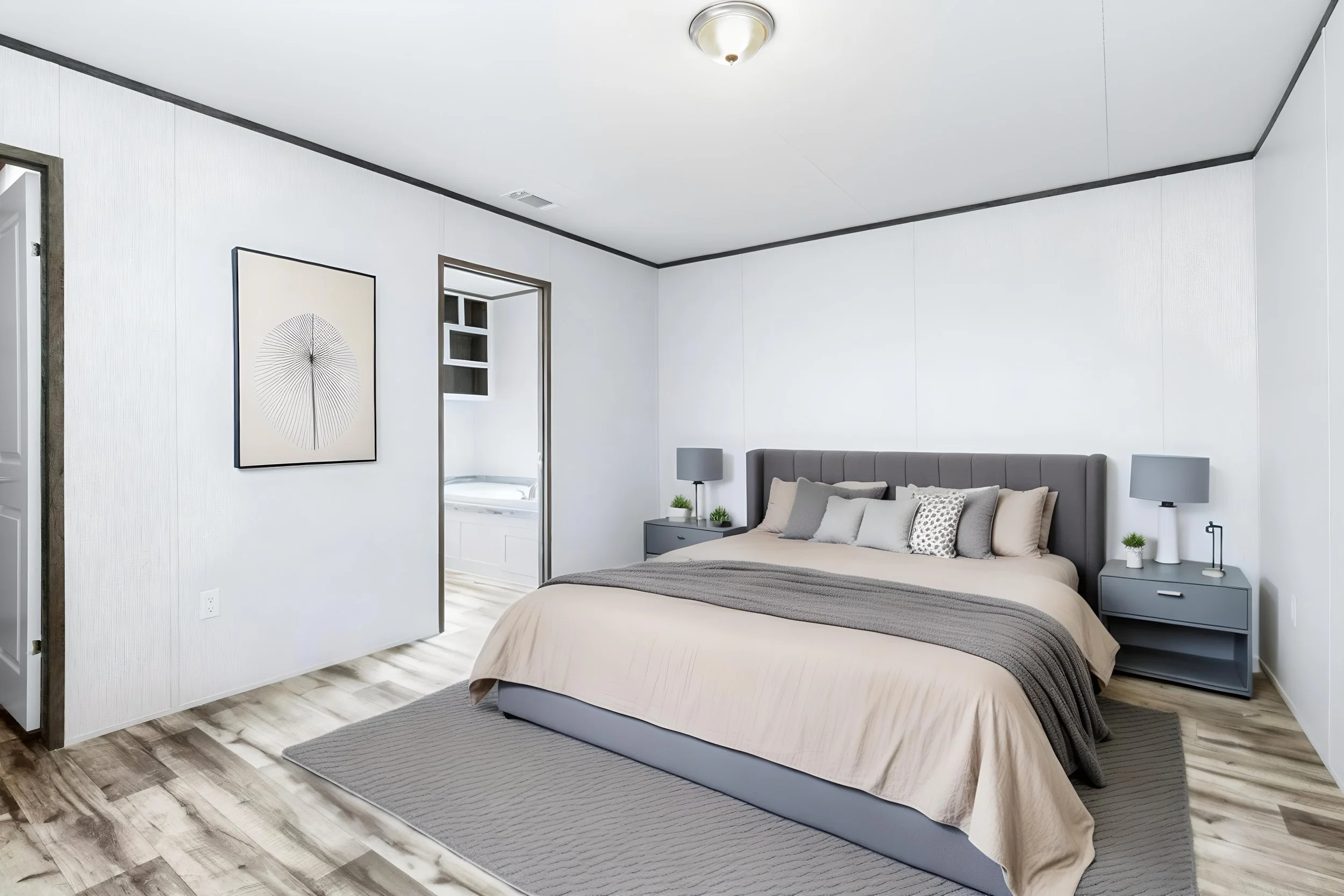 Minimalist bedroom with a queen-sized bed, gray headboard, beige bedding, and gray side tables with lamps. Light wood flooring and white walls with black trim, artwork on the wall, and a small window.