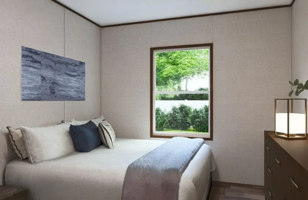 A bedroom with beige walls, a window showing greenery outside, a bed with white and dark blue pillows, a blue throw blanket, a piece of abstract art on the wall, and a brown dresser with a lamp and a plant.