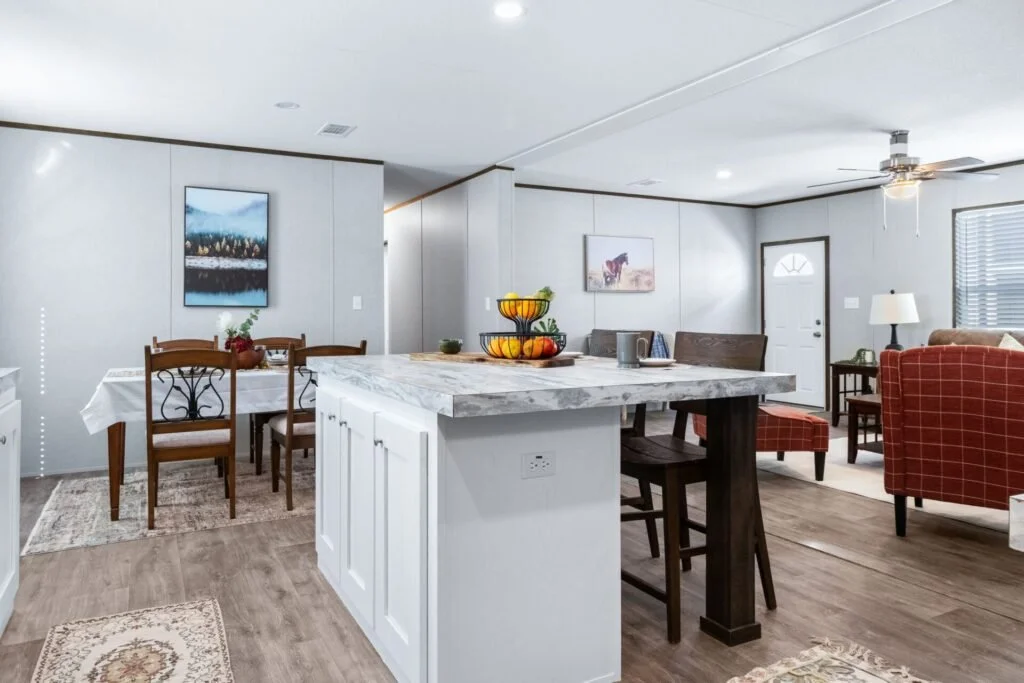 Open-concept living and dining area with a kitchen island, white walls, wooden flooring, and furniture including a dining table, chairs, armchairs, and a side table with a lamp.