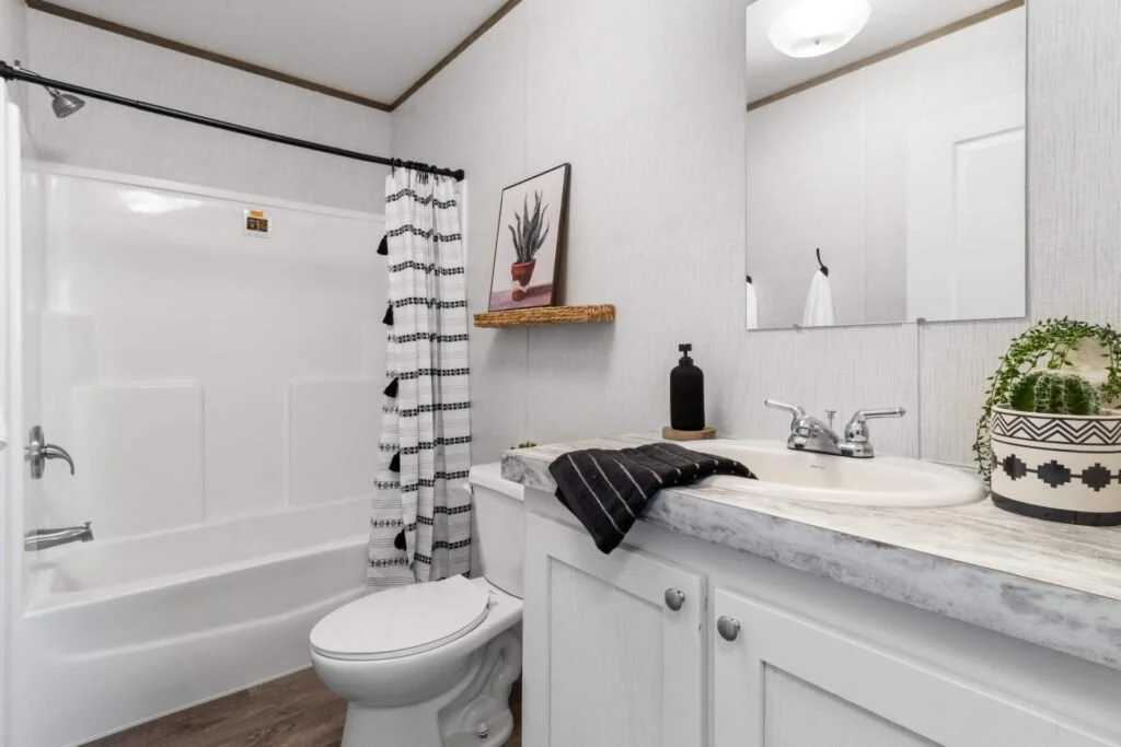 A small bathroom with a white bathtub, black and white shower curtain, white toilet, and a white vanity with a marble countertop. There is a large mirror above the sink, a black soap dispenser, a potted plant, and framed wall art of a cactus on a flo