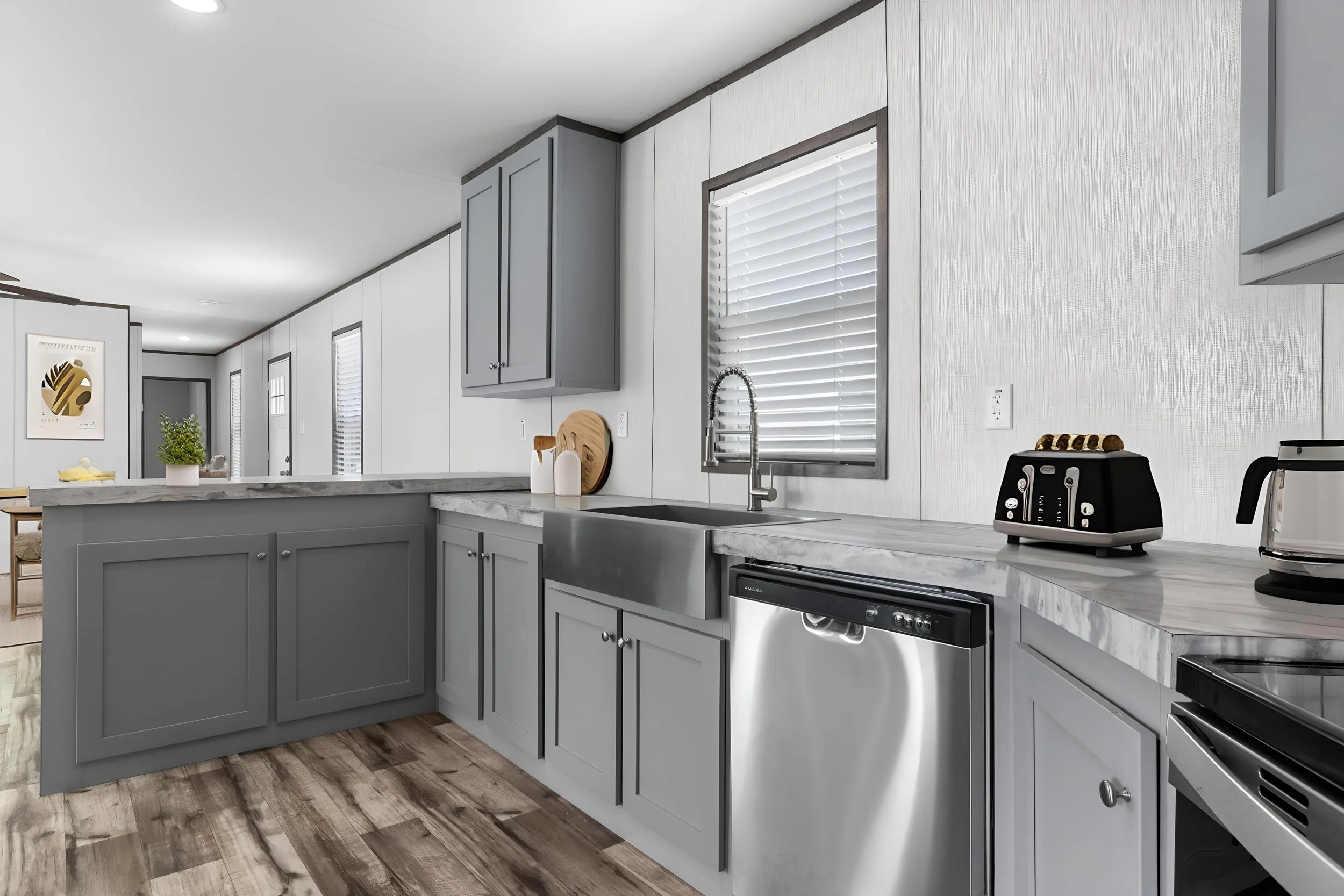 Modern kitchen with gray cabinets, white countertops, a stainless steel sink, window with blinds, toaster, coffee maker, and wooden flooring.