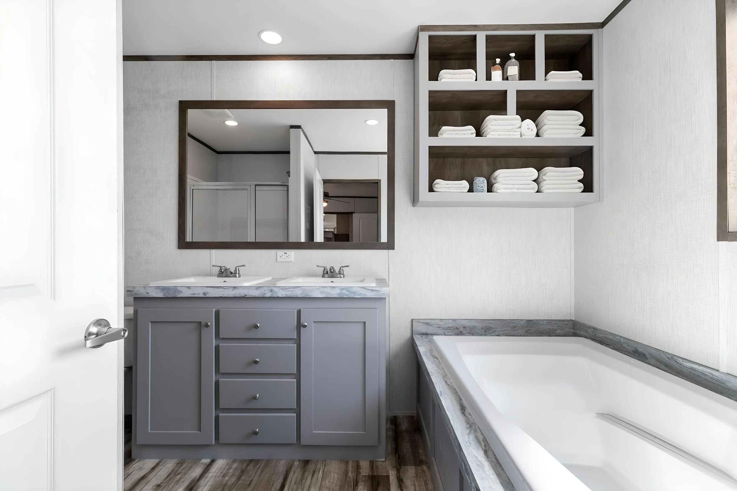 A bathroom featuring a double sink vanity with a marble countertop, a mirror, open shelves with towels and toiletries, a bathtub with marble surround, and wood-look flooring.