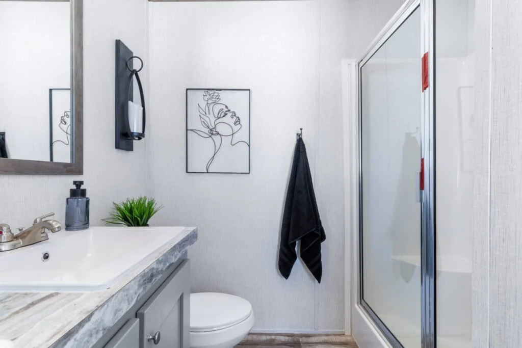 Bathroom with a sink, mirror, soap dispenser, small plant, framed line art of a woman, towel hanging on a hook, and a shower with a sliding door.