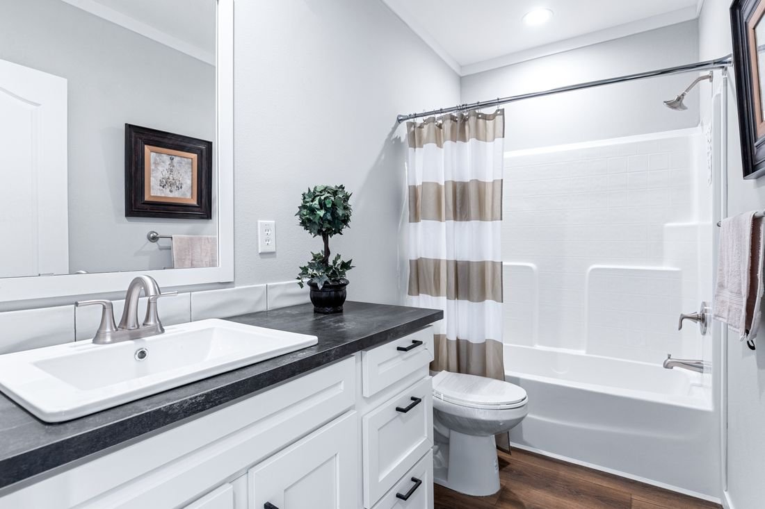 Bathroom with white cabinets, a black countertop, a sink, a potted plant, a mirror, a framed picture, a towel, a toilet, a bathtub with a striped shower curtain, and hardware fixtures.