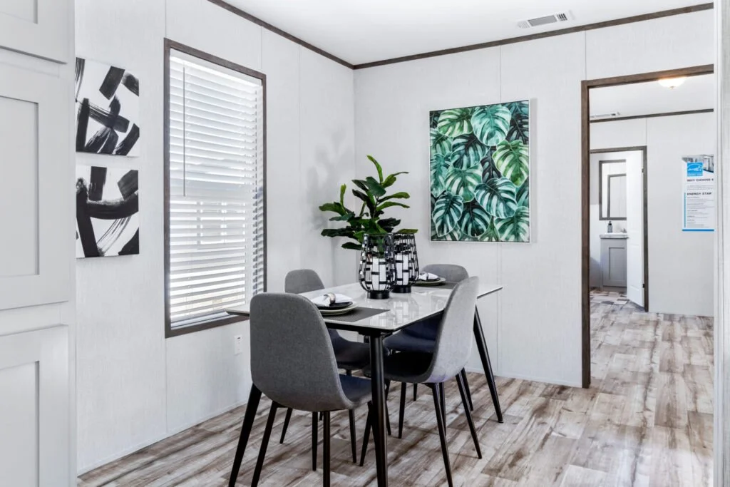 A dining room with a rectangular table, surrounding four gray cushioned chairs, a large potted plant in a black and white vase, and two pieces of wall art featuring black and white abstracts and green tropical leaves.