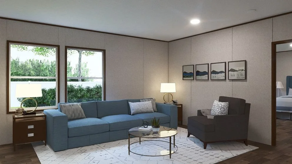Living room with blue sofa, gray armchair, two side tables with lamps, framed artwork on walls, large windows with greenery outside, and a partially visible bedroom.