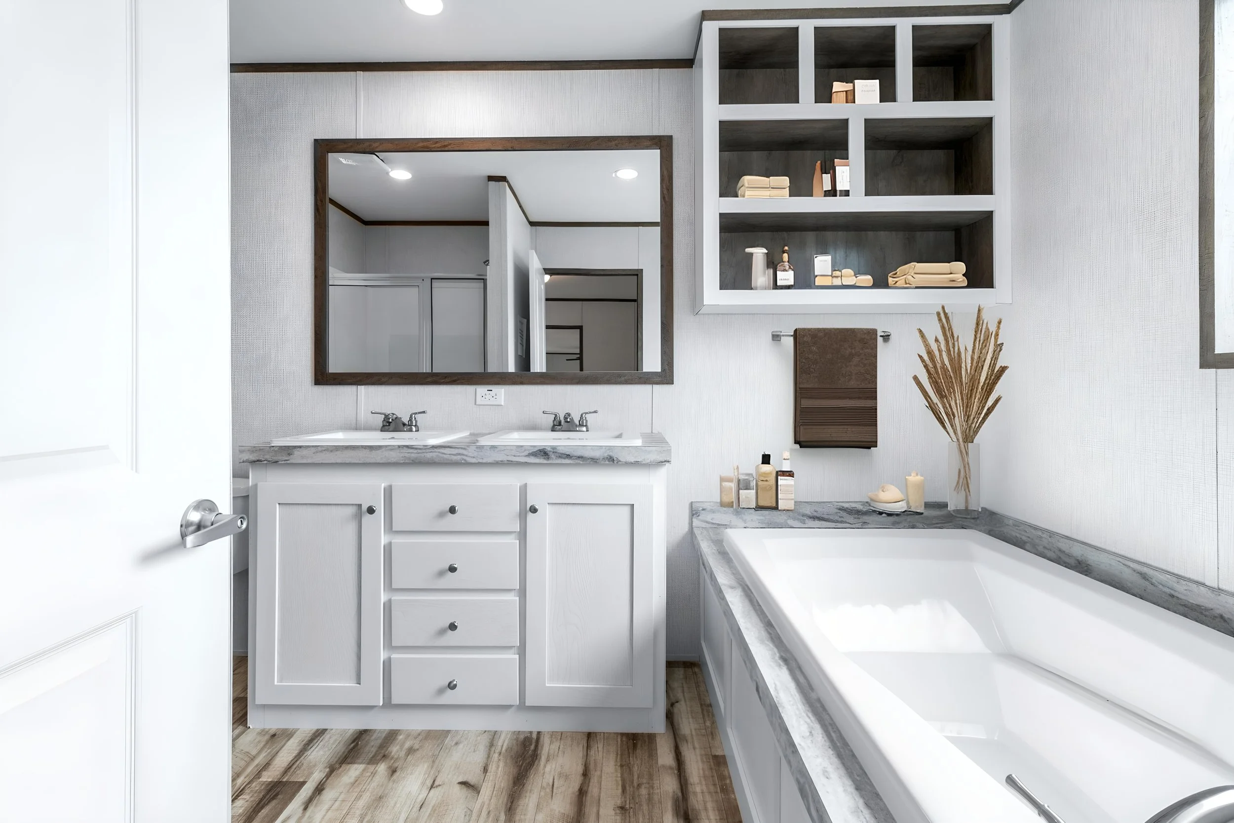 Modern bathroom with white vanity, dual sinks, large mirror, open shelves with storage boxes, and a bathtub with gray marble surround, wood flooring, and a vase with dried plants.