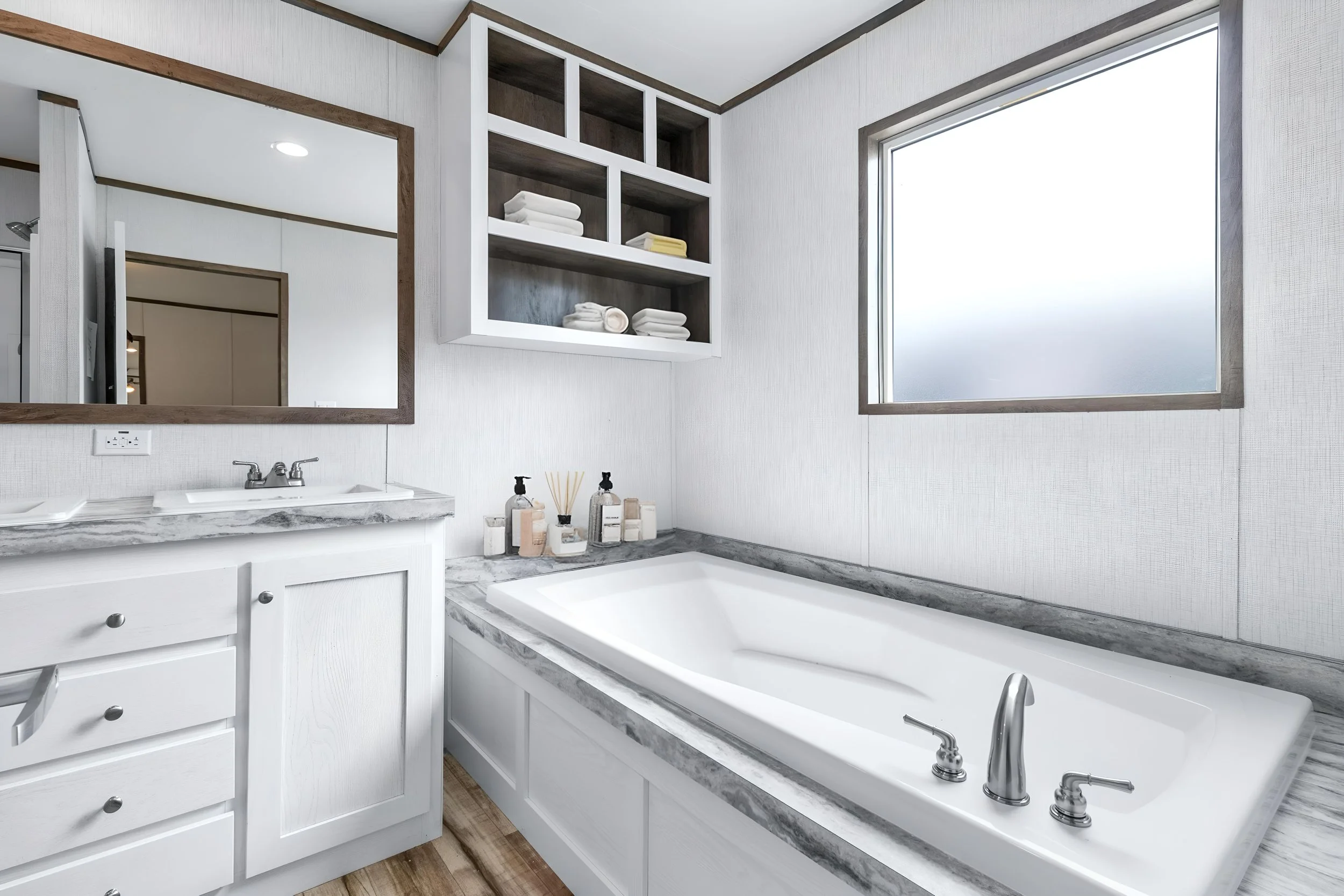 Bathroom with a white vanity, marble countertop, large mirror, built-in bathtub with marble surround, open corner shelving with towels and folded linens, and a window with frosted glass.