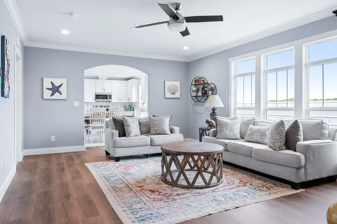 Bright living room with light gray walls, large windows, two white sofas with patterned pillows, a round wooden coffee table, a side table with a lamp, and artwork on the walls. The room opens to a kitchen with white cabinets and dining area.