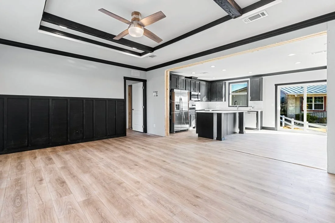 Open-concept living space with light wood flooring, black wainscoting, and a kitchen with black cabinets, stainless steel appliances, and a large window. Recessed lighting and a ceiling fan are visible.