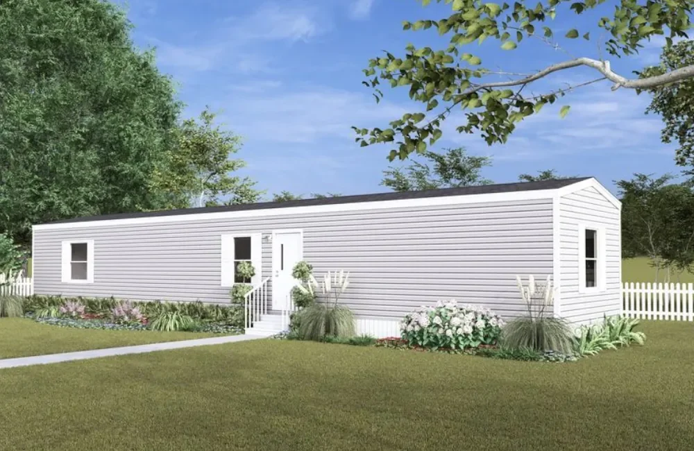 A white manufactured home with a black shingle roof, situated in a landscaped yard with grass, bushes, and trees, and a white picket fence in the background.