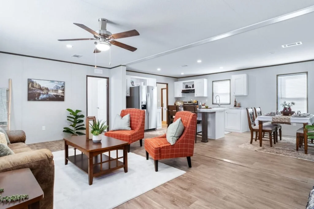 Open-concept living room and kitchen with white cabinets, wooden dining table, and orange accent chairs.