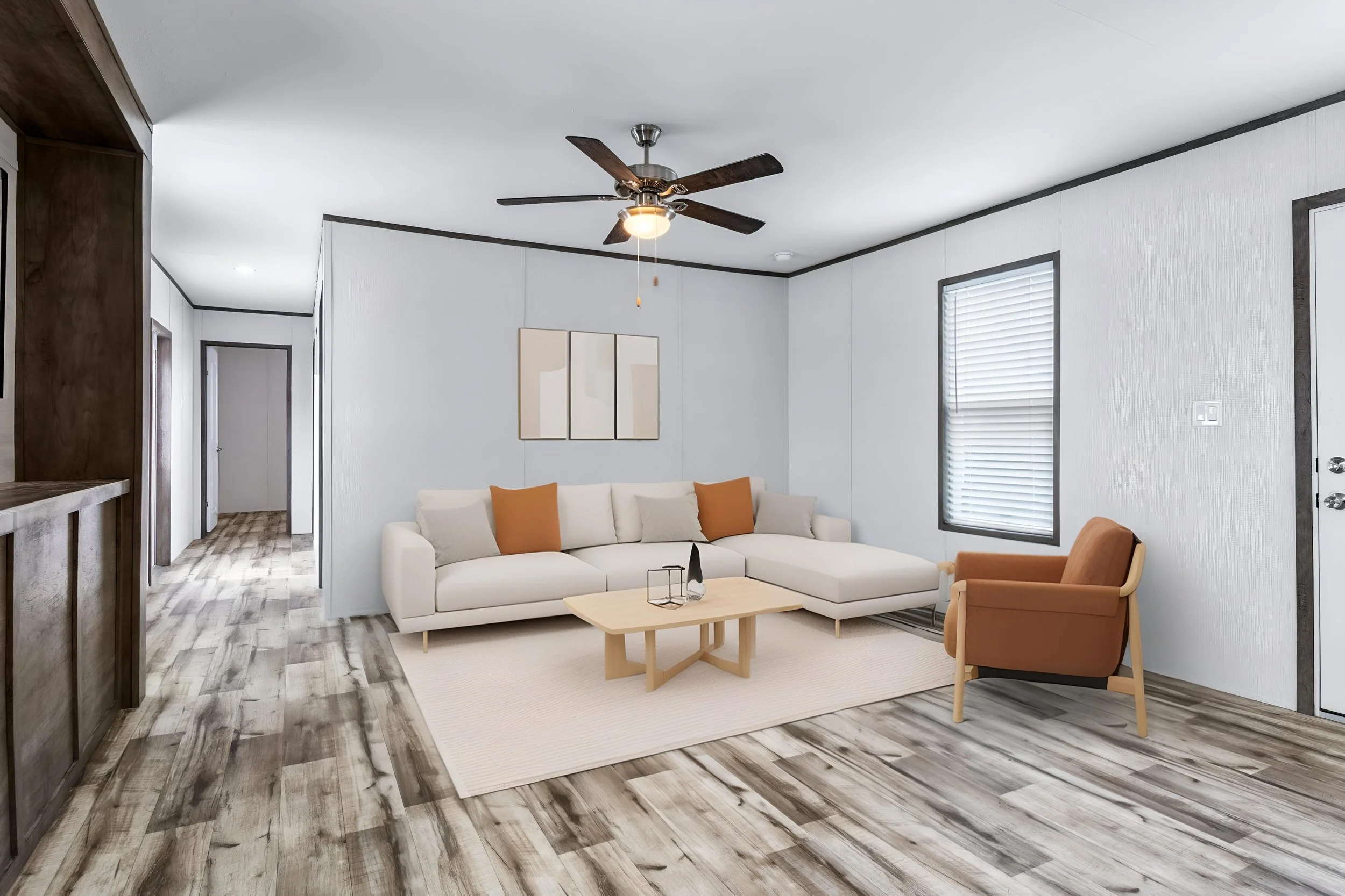 Living room with white sectional sofa, brown armchair, wooden coffee table, artwork on wall, ceiling fan, window with blinds, and wood-like flooring.