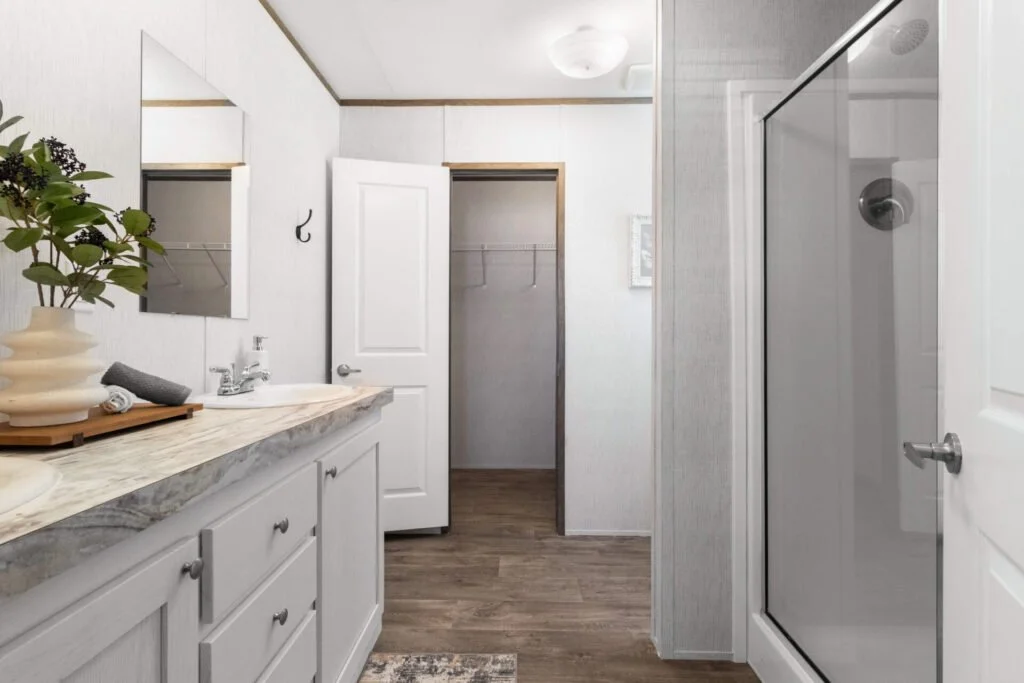 Bathroom with a marble countertop, sink, mirror, and a houseplant. Open closet with wire shelf, glass shower door, and wood flooring.