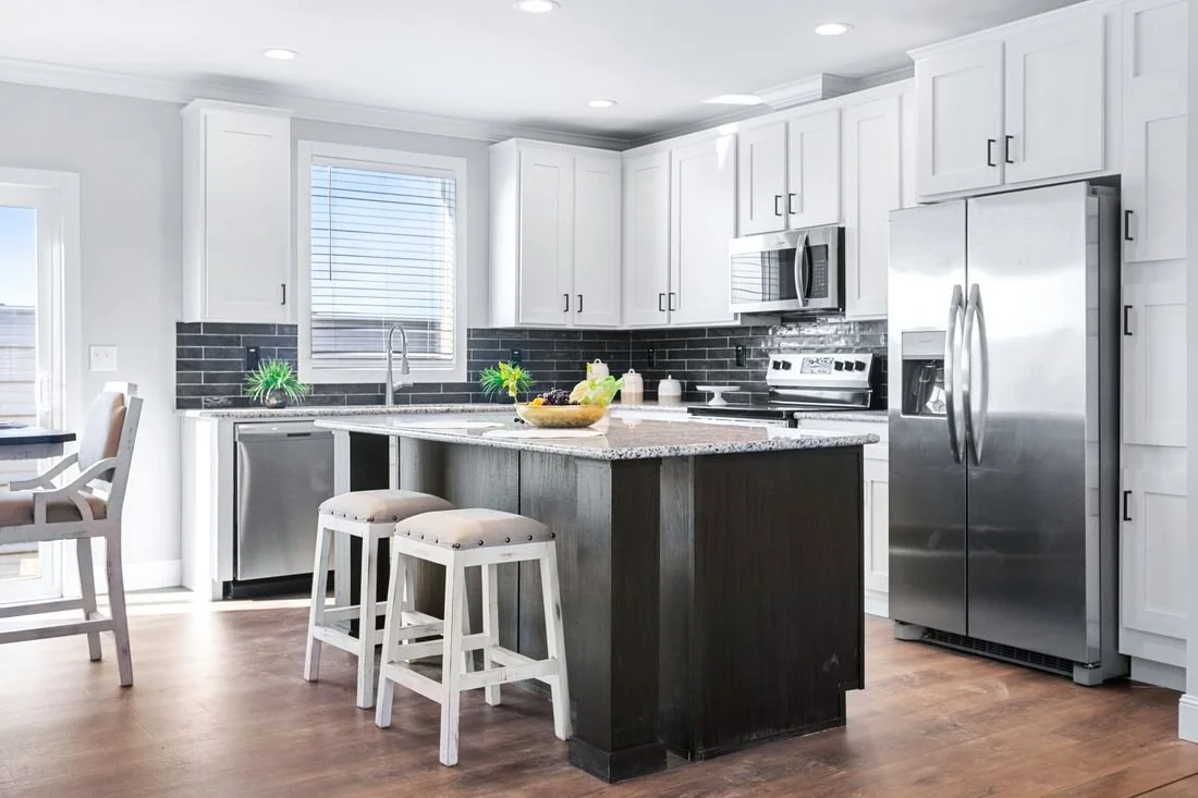 Modern kitchen with white cabinets, black backsplash, and stainless steel appliances including a refrigerator, microwave, and oven. A kitchen island with a granite countertop has two white barstools, and there is a window with blinds above the sink. 