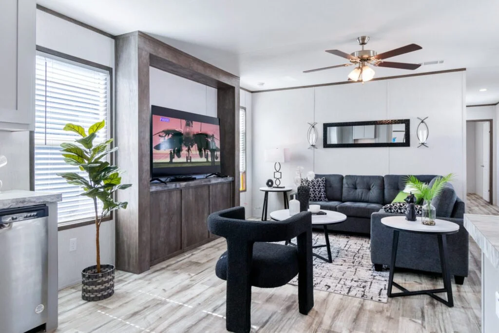 Living room with gray couch, black chairs, a TV on a wooden stand, a ceiling fan, a mirror, and decorative plants.