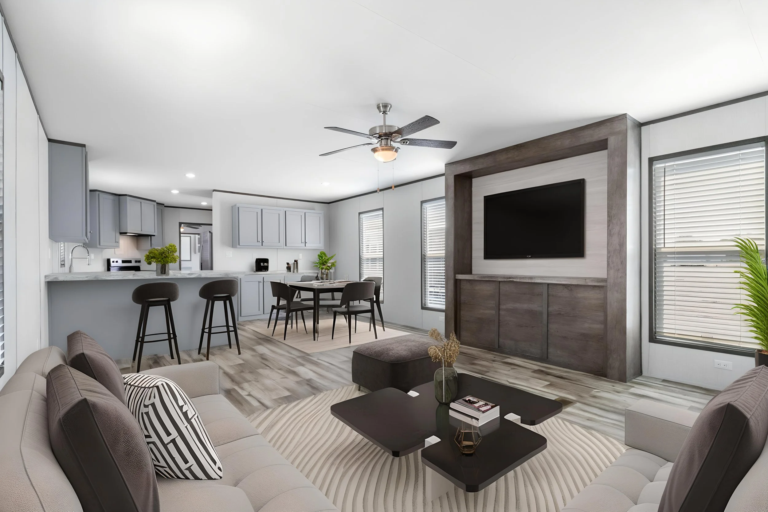 Open-concept living room and kitchen with modern decor, gray cabinets, wooden flooring, a ceiling fan, TV mounted on a wooden accent wall, and a large window with blinds.