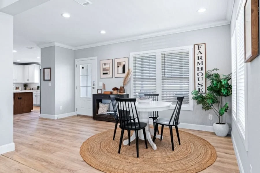 A bright dining area with a round white table and four black chairs on a circular jute rug, next to a large window with white blinds. A tall potted plant is in one corner and a sign that reads 'FARMHOUSE' is on the wall. The background shows part of 