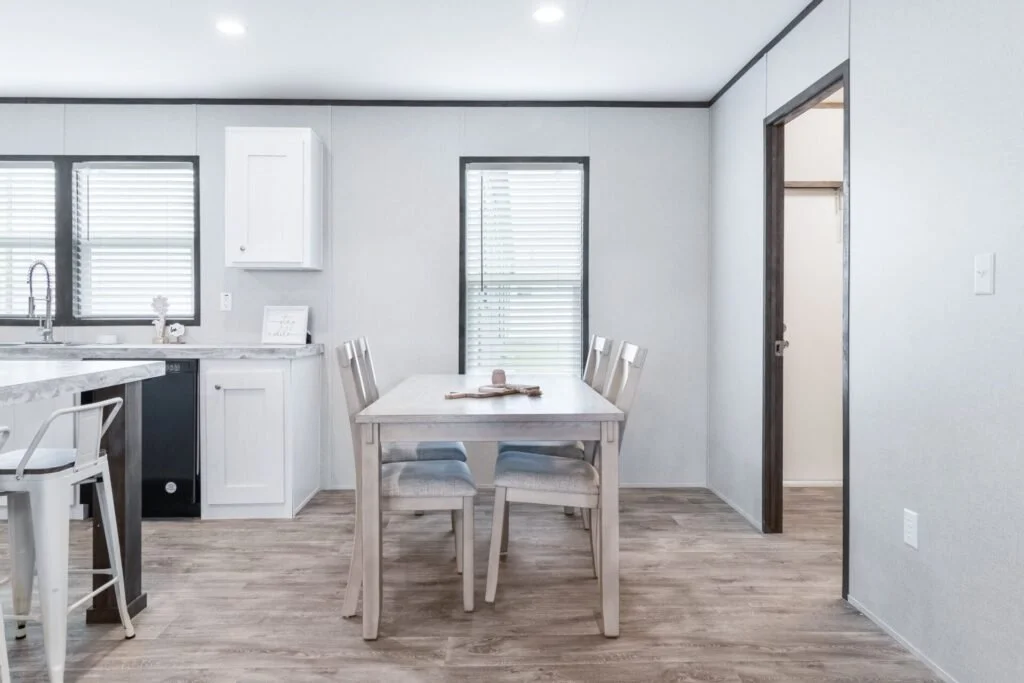 A minimalist dining area with a wooden table, four chairs, two windows with blinds, and a doorway leading to another room.