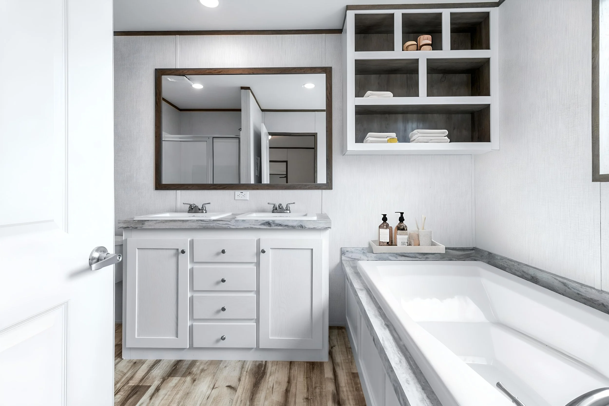 A bathroom with a white vanity, double sinks, a large mirror, shelves with folded towels, and a bathtub with a marble surround.