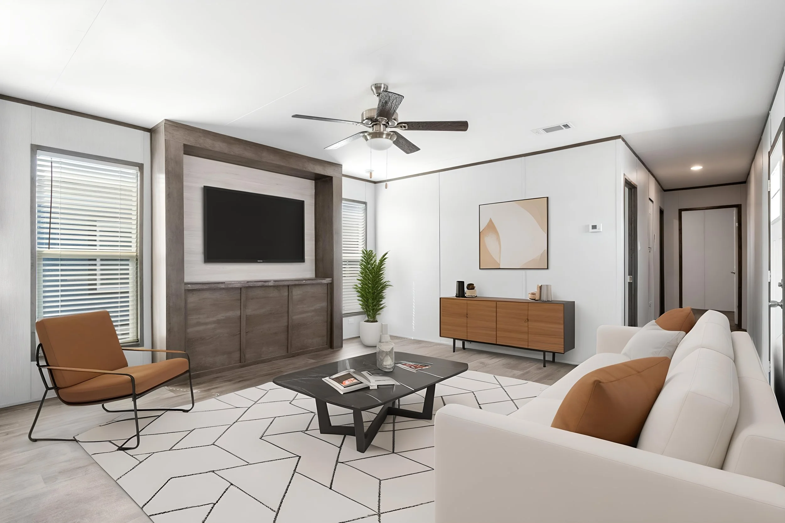 Living room with white walls, large windows with blinds, a ceiling fan, a flat-screen TV, a wooden sideboard with decor, a modern geometric rug, a white sofa with tan cushions, a brown armchair, a coffee table with magazines, and abstract wall art.