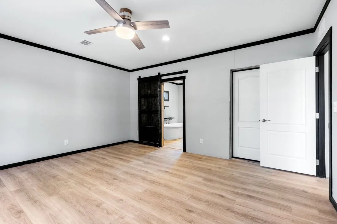 Empty bedroom with light wood flooring, white walls with black trim, a ceiling fan, and an open door leading to a bathroom with a bathtub.