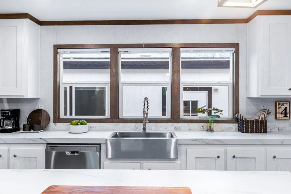 Modern kitchen with white cabinets, a large farmhouse sink, and a window above it, decorated with a potted plant, a tray with a drink, and a sign that says 'Party of 4'.