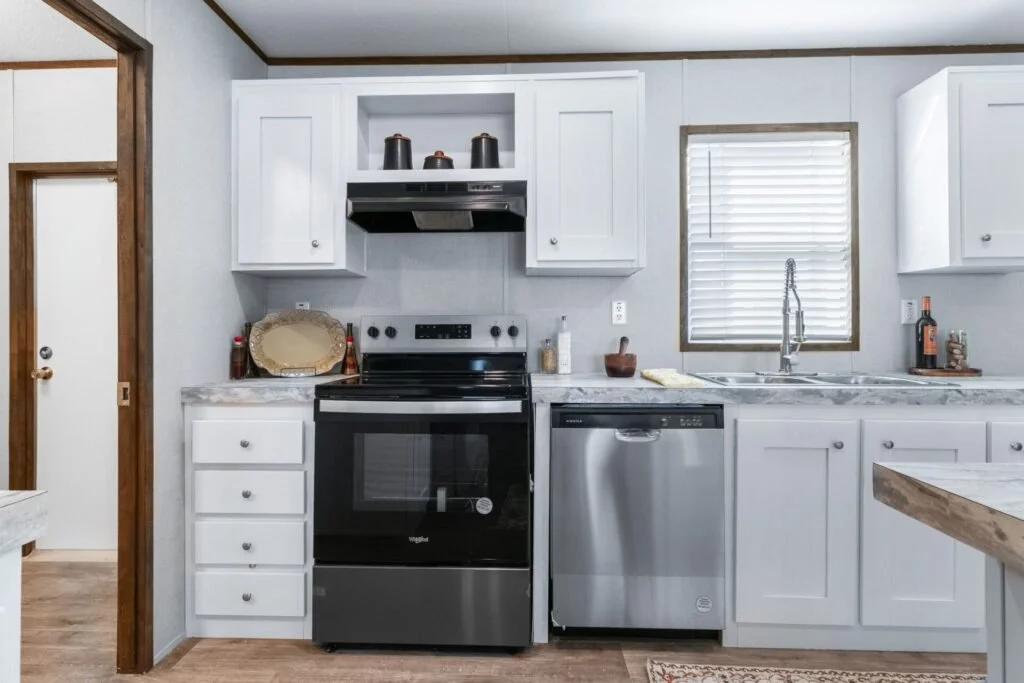 Modern kitchen with white cabinets, stainless steel oven and dishwasher, marble countertops, and a window with blinds above the sink.