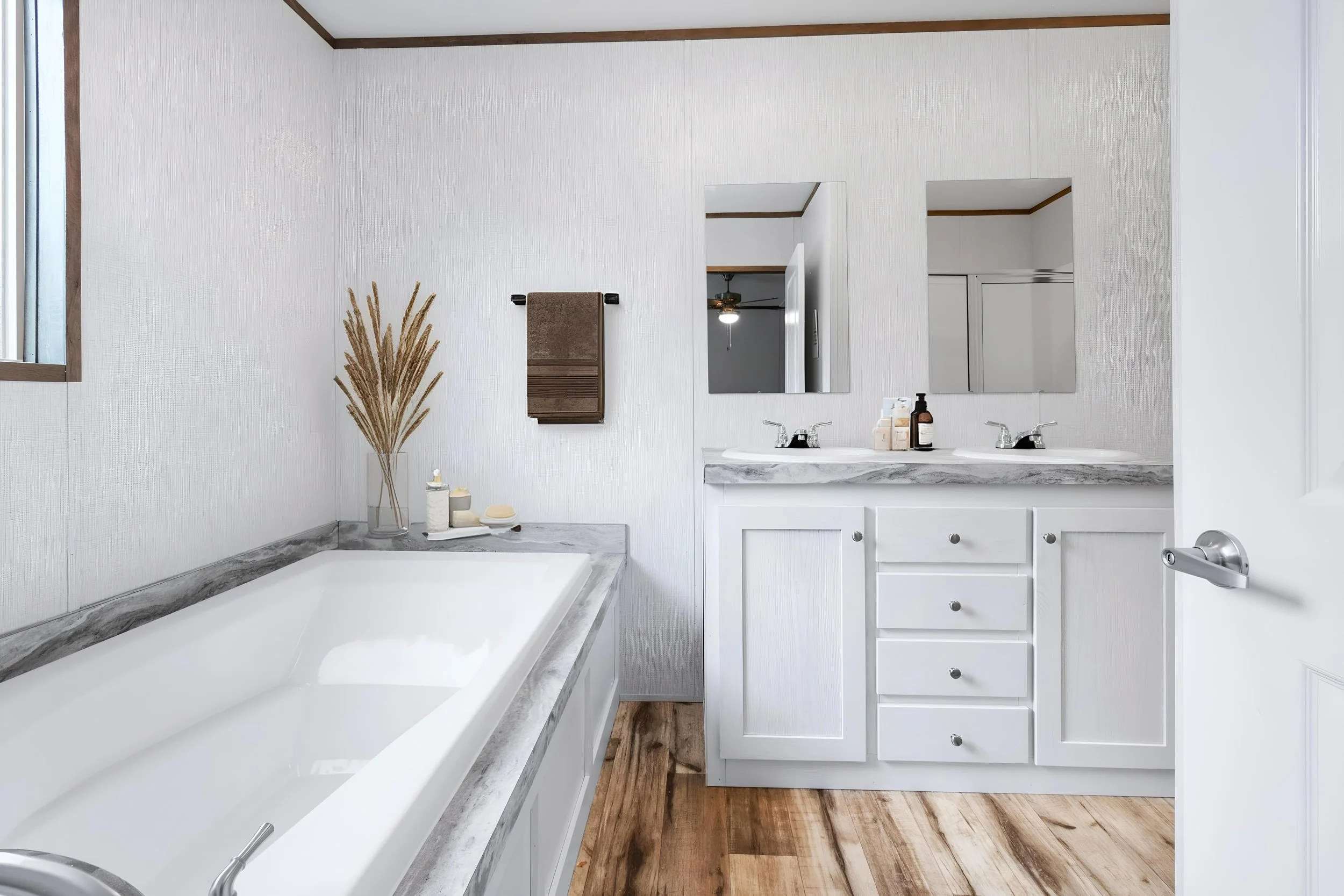 Bathroom with a white vanity, two sinks, and a large mirror. The corner shows a bathtub with a marble surround. Decorative dried plants and toiletries are on the marble ledge. A small cabinet is mounted on the wall, and a window brings natural light 